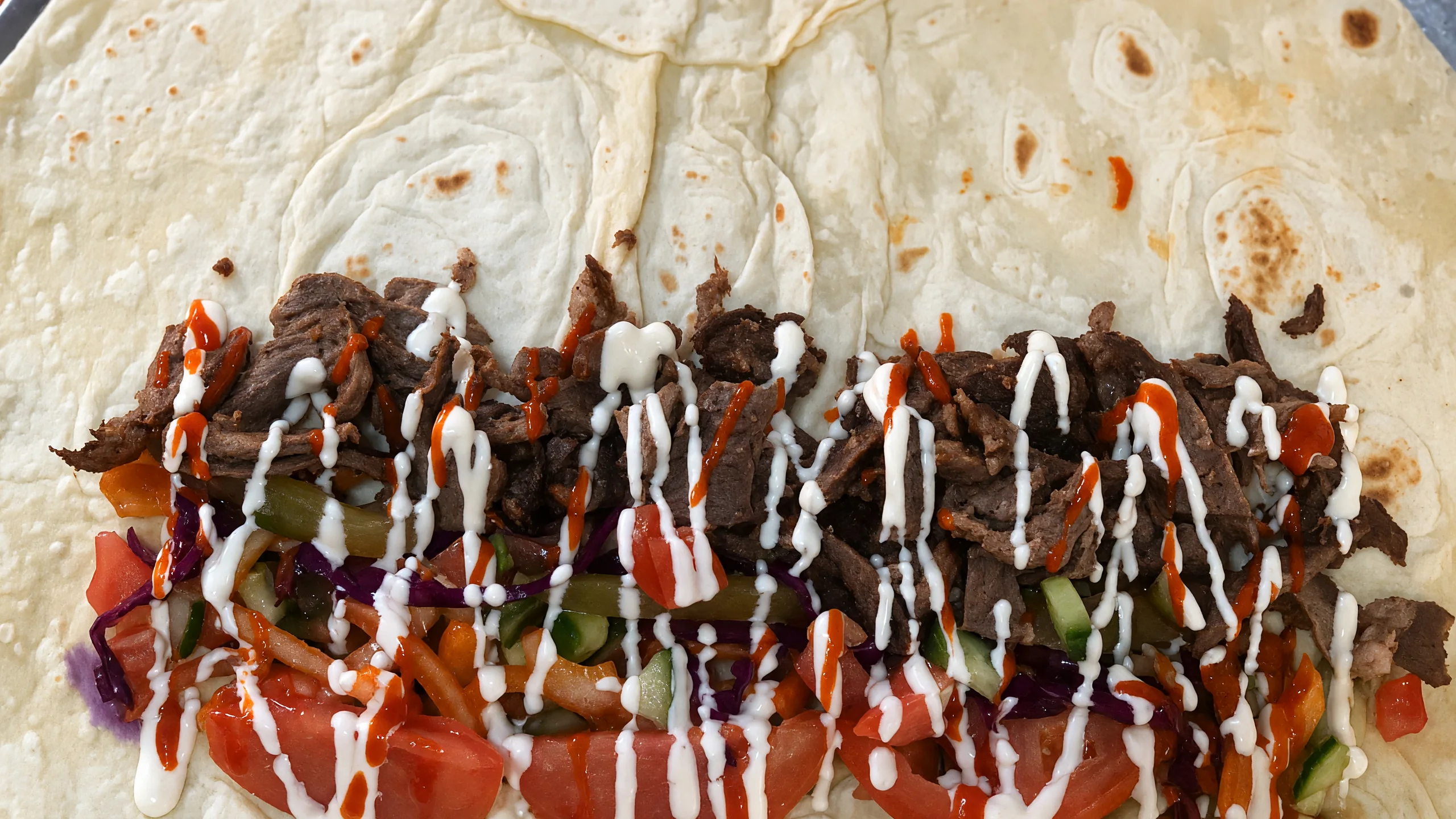 Beef shawarma wrap being built on fresh lavash with tomato, pickles, garlic sauce and hot sauce
